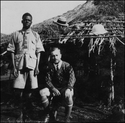 Eric Beavon sitting in front of a hut with an assistant